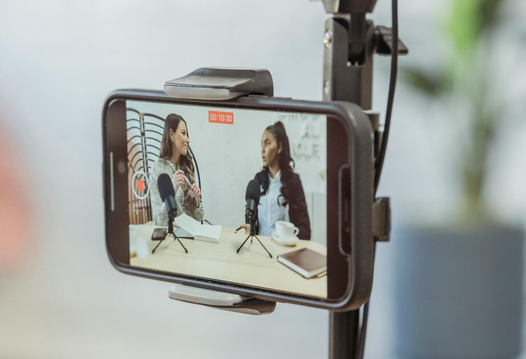 Young diverse women sitting at table with microphones and gadgets and talking while recording podcast on smartphone placed on tripod in studio